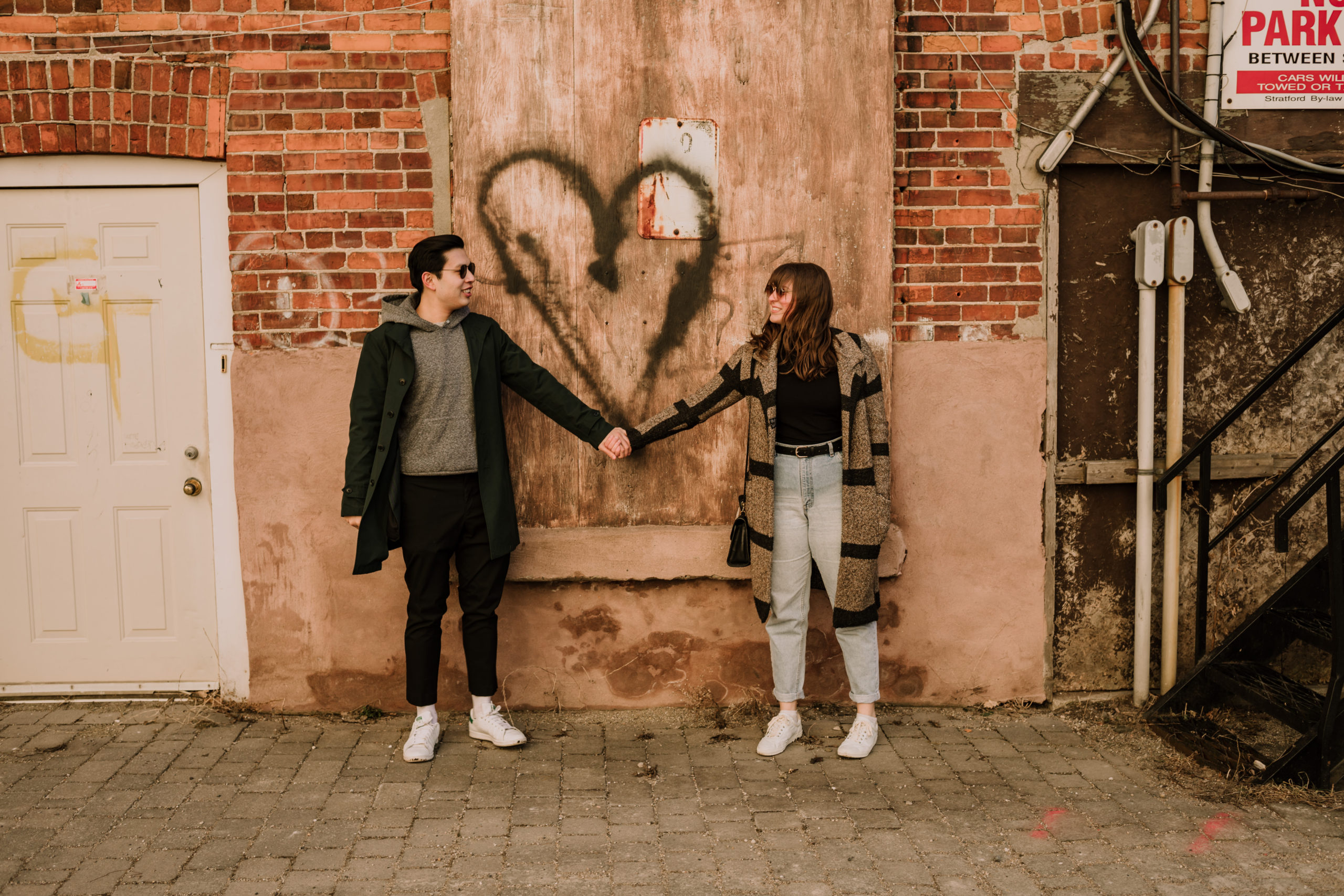 Couple hold hands in front of a graffiti heart during a photo session with Cara Gilhula Photography in Stratford, Ontario